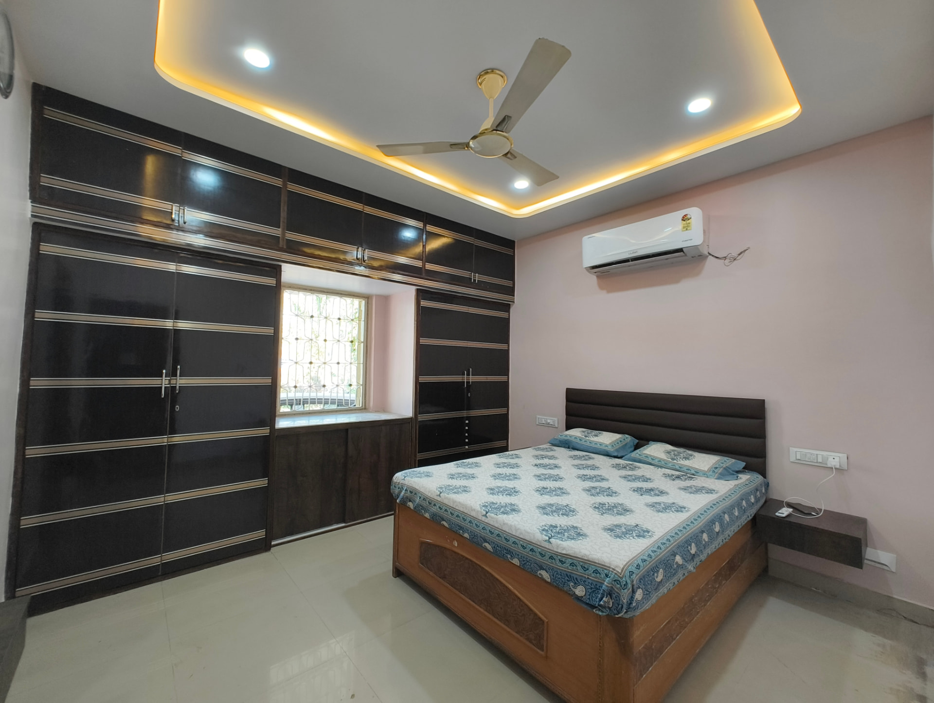 Master bedroom by Gharva Studio with dark wood wardrobes inlaid with gold stripes, a warm-lit tray ceiling and a blush accent wall